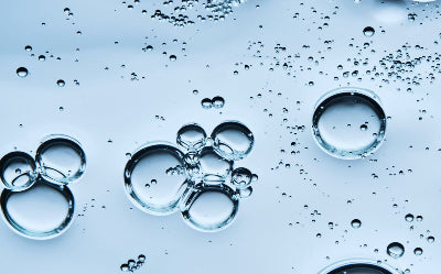 Une vue macroscopique montrant des bulles et des gouttes transparentes de différentes tailles flottant dans un liquide bleu clair et pur. L'ensemble évoque une structure moléculaire fluide et rafraîchissante, avec des jeux de lumière et de reflets sur les parois des sphères.